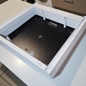 Puede incluir: Un marco cuadrado blanco xTool con una placa base interior negra. El marco cuenta con pilares de soporte verticales blancos en las esquinas y puntos centrales. El texto XTOOL est&aacute; impreso en blanco sobre la base negra, junto con marcas de cruz blancas y las etiquetas AV-G, AV y AV-D impresas en los bordes del marco blanco.
