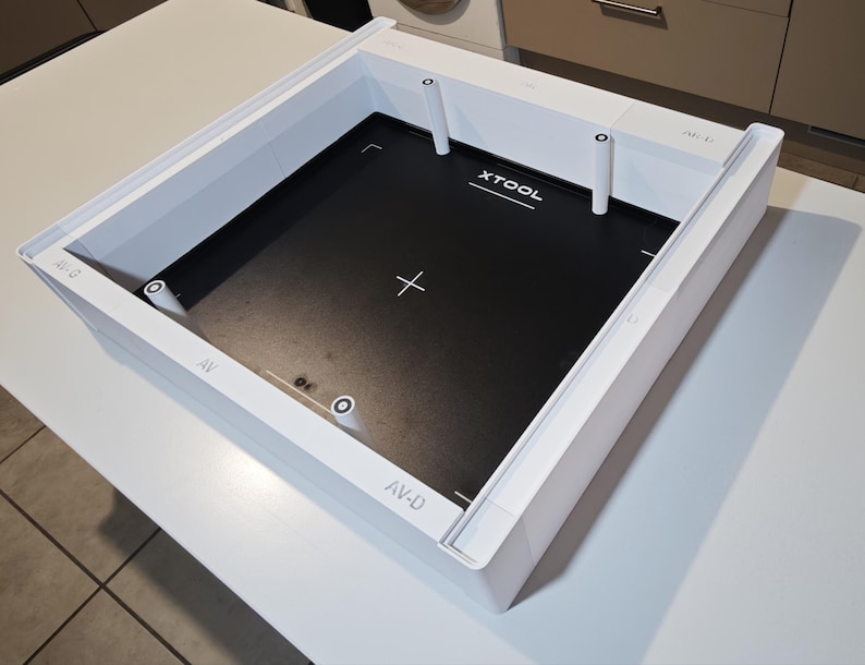 Puede incluir: Un marco cuadrado blanco xTool con una placa base interior negra. El marco cuenta con pilares de soporte verticales blancos en las esquinas y puntos centrales. El texto XTOOL est&aacute; impreso en blanco sobre la base negra, junto con marcas de cruz blancas y las etiquetas AV-G, AV y AV-D impresas en los bordes del marco blanco.