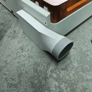 May include: A white laser cutter machine featuring a large, translucent amber-colored lid. Attached to the side of the white base is a white, rectangular exhaust vent adapter that transitions into a circular gray opening for hose attachment.