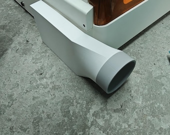 Puede incluir: Una máquina de corte láser blanca con una tapa grande y translúcida de color ámbar. En el lateral de la base blanca hay un adaptador de ventilación rectangular blanco que termina en una abertura circular gris para manguera.