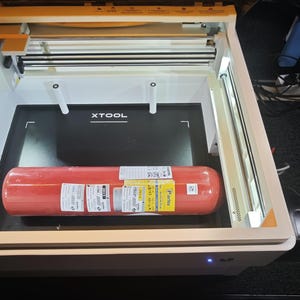 Puede incluir: Una m&aacute;quina de corte l&aacute;ser xTool con marco blanco e iluminaci&oacute;n interna, que presenta una superficie de trabajo negra con el logotipo xTool. Un extintor cil&iacute;ndrico rojo con etiquetas de env&iacute;o blancas est&aacute; colocado en la base.