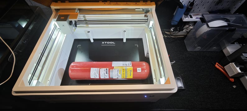 Puede incluir: Una m&aacute;quina de corte l&aacute;ser xTool con marco blanco e iluminaci&oacute;n interna, que presenta una superficie de trabajo negra con el logotipo xTool. Un extintor cil&iacute;ndrico rojo con etiquetas de env&iacute;o blancas est&aacute; colocado en la base.