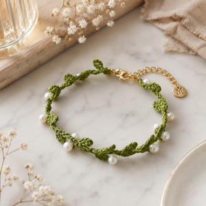 May include: A green crocheted bracelet featuring a vine-like pattern with small white pearls interspersed throughout. The bracelet includes a gold-toned metal lobster clasp, an adjustable chain, and a small circular charm engraved with the letter M.