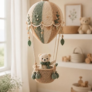 May include: A crocheted hot air balloon mobile featuring a beige and sage green striped balloon canopy with embroidered leaf details and hanging tassels. A small crocheted teddy bear wearing a green scarf sits inside the woven basket.