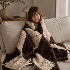 May include: A crochet blanket featuring a geometric diamond pattern in alternating dark brown and cream yarn. The textile displays a textured stitch construction with openwork detailing along the edges of each diamond shape, creating a repeating motif.