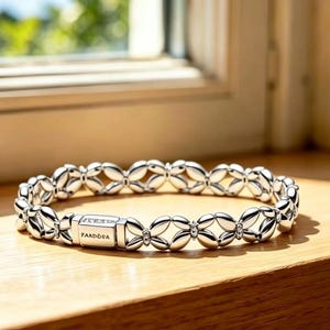 May include: A silver-toned bracelet featuring a repeating pattern of interconnected petal-like shapes. The piece includes a rectangular clasp engraved with the word PANDORA, providing a secure closure for this polished metal jewellery item.