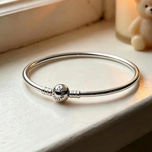 May include: A polished silver bangle bracelet featuring a round, spherical clasp engraved with the word Pandora. The bracelet has a smooth, circular band design with two small ridges on either side of the central clasp mechanism.