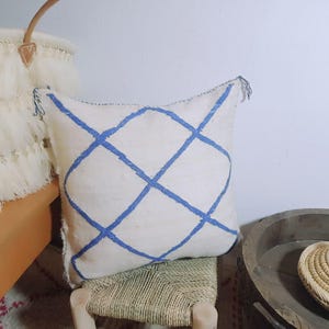 Puede incluir: Un cojín cuadrado de color blanco roto con un patrón de rombos bordado en azul. El cojín tiene pequeñas borlas azules en las esquinas superiores y una superficie de tela texturizada. El diseño consiste en líneas azules que se cruzan.