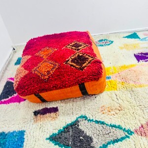 May include: A square floor pouf featuring a deep red, textured top surface with orange and dark brown diamond patterns. The sides of the pouf are a solid orange fabric with black vertical accents, resting on a cream rug with colorful geometric shapes.