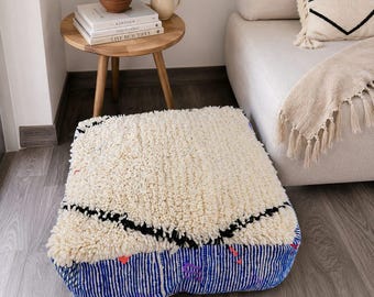 May include: A square floor cushion featuring a thick, cream-colored shaggy wool top with a black geometric line pattern. The sides are constructed from blue fabric with white vertical stripes, accented by small, scattered red and purple details.