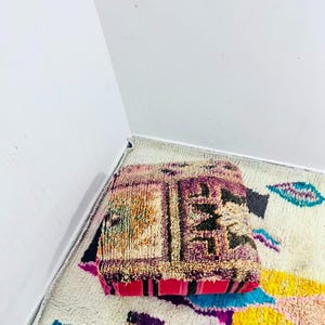May include: A square floor cushion featuring a textured, hand-knotted wool surface with a vibrant color palette of purple, pink, orange, and black geometric patterns. The sides of the cushion are accented with bold, solid pink and black vertical stripes.