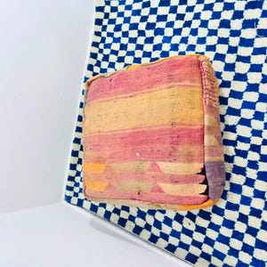 May include: A square floor pouf featuring a woven textile surface with horizontal stripes in shades of pink, orange, and yellow. The fabric displays a subtle geometric pattern along the bottom edge and a textured, multi-colored side panel.