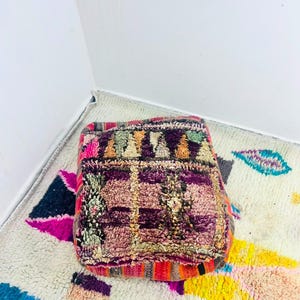 Puede incluir: Un puf de suelo cuadrado con una superficie texturizada anudada a mano en una vibrante paleta de colores que incluye morado intenso, magenta, naranja y verde suave. La superficie muestra un patrón geométrico con rayas horizontales.
