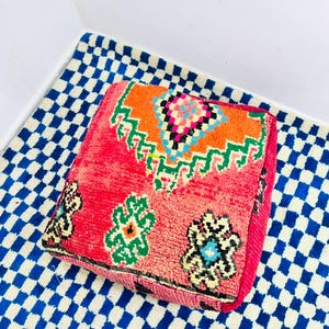Könnte beinhalten: Ein quadratisches Bodenkissen mit einer leuchtend roten Basis und einem großen geometrischen Muster in Orange, Grün und Blau auf der Oberseite. Die Seiten sind mit kleineren, komplexen blumenartigen Motiven in Schwarz, Grün und Gelb verziert.