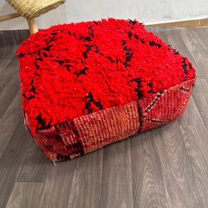 Peut inclure: Un pouf de sol carré doté d'un dessus en poils longs et épais de couleur rouge avec des accents noirs. Les côtés du pouf sont constitués d'un textile tissé plus plat dans des tons rouge et orange, présentant un motif géométrique linéaire.