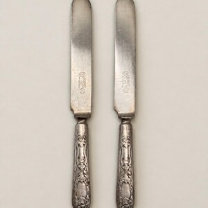 May include: Two silver-toned metal dinner knives with rounded blades and ornate, embossed floral handles. Each blade features a stamped mark that reads Rogers 1881. The knives have a metallic finish with visible signs of age and patina.