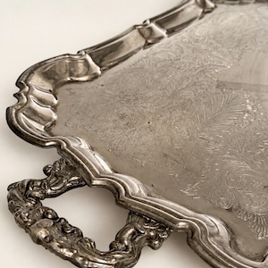 May include: A silver-toned metal serving tray featuring an intricate engraved floral pattern on its surface. The tray has a scalloped, raised edge and two ornate, textured handles on the sides, showcasing a metallic finish throughout.
