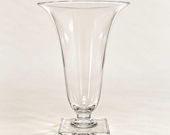 May include: A clear glass vase featuring a wide, flared opening that tapers down to a slender neck, supported by a sturdy, square-shaped base. The transparent vessel has a smooth, polished surface and a symmetrical design, standing upright.