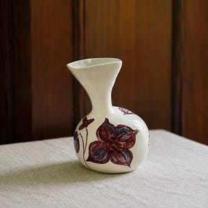 May include: A cream-colored ceramic vase with a rounded base and a flared neck. The surface is decorated with hand-painted dark reddish-brown floral motifs featuring blue-toned shading on the petals, arranged around the bulbous body of the vessel.