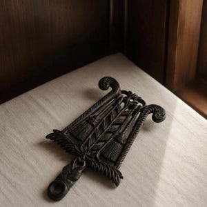 May include: A dark brown cast iron trivet featuring a decorative openwork design. The pattern includes two upright broom motifs flanked by wheat stalks, with curled scrollwork at the top and a small circular handle extension at the bottom.