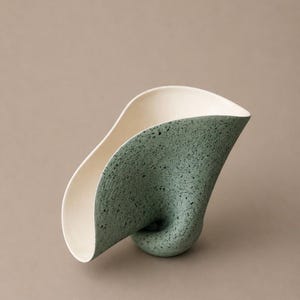 May include: A sculptural ceramic vessel with an organic, curved form. The exterior features a matte, sage green finish with dark speckles, while the interior is a smooth, solid off-white. The base is tapered and twisted into a singular point.