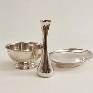May include: A set of three silver-toned metal items consisting of a small bowl with a pedestal base, a tall slender bud vase with a flared rim, and a shallow circular dish. Each piece features a polished, reflective surface and smooth curves.