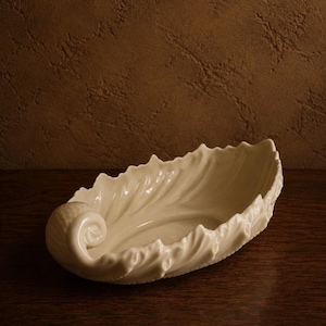 May include: An off-white ceramic bowl shaped like a curled leaf with a prominent spiral at one end. The bowl features intricate, raised vein-like textures along its edges, mimicking the natural appearance of a foliage element in a glossy finish.