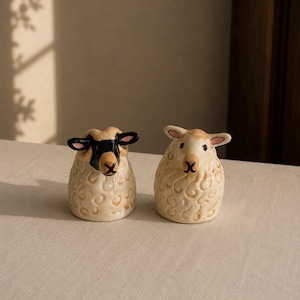 May include: Two ceramic sheep figurines with cream-colored, textured wool bodies. The left sheep has a black face and ears with curled horns, while the right sheep has a light tan face and pink-lined ears. Both have small, dark X-shaped mouths.