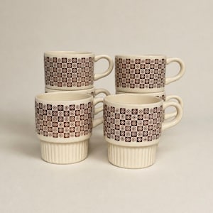 May include: A set of four cream-colored ceramic mugs stacked in two columns. Each mug features a brown checkered pattern with small floral icons inside the squares and a ribbed base design with a curved handle on the side of each piece.