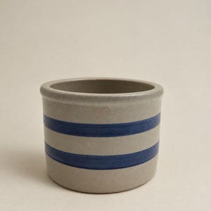 May include: A cylindrical ceramic crock with a smooth, light gray finish and two horizontal dark blue stripes encircling the exterior. The vessel features a thick, rounded rim and a flat base, presenting a simple, functional design for storage.