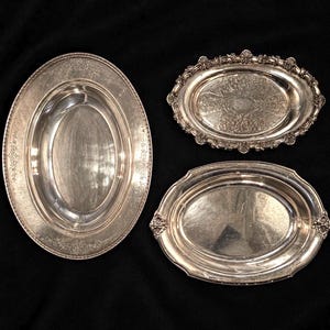 May include: Three oval silver-toned serving trays with metallic finishes. The largest tray features a simple beaded rim. The medium tray has an ornate, scalloped edge with intricate floral engravings. The third tray has a smooth, polished rim.