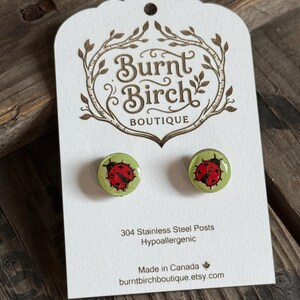 May include: A pair of round stud earrings featuring a red and black ladybug graphic on a light green background. The earrings are mounted on a white display card with the text Burnt Birch Boutique, 304 Stainless Steel Posts, Hypoallergenic.