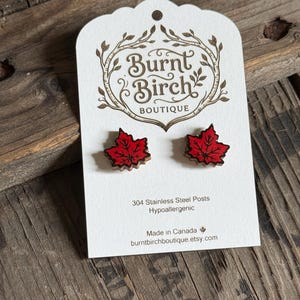 Peut inclure: Une paire de boucles d'oreilles clous en forme de feuille d'érable rouge sur un carton de présentation blanc avec le texte Burnt Birch Boutique, 304 Stainless Steel Posts, Hypoallergenic, Made in Canada et burntbirchboutique.etsy.com.