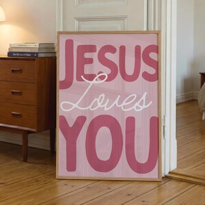Puede incluir: Un estampado rosa enmarcado con las palabras "JESUS Loves YOU" en una fuente audaz y juguetona. El texto está en tonos de rosa y blanco, sobre un fondo rosa claro. El marco es de color madera natural.