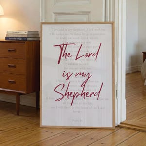 May include: A framed print with the text "The Lord is my Shepherd" in a burgundy script font. The background is a light gray with printed verses. The frame is a light brown wood.