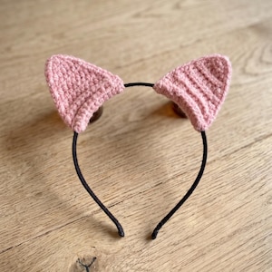 May include: A thin black headband featuring two triangular, light pink crocheted cat ears attached to the top. The ears have a visible textured stitch pattern and are positioned symmetrically on the band, creating a simple and functional accessory.