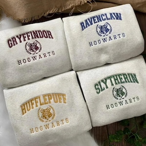 May include: Four light grey sweatshirts featuring embroidered text and crests for the four Hogwarts houses. The top left displays Gryffindor in maroon, top right Ravenclaw in blue, bottom left Hufflepuff in gold, and bottom right Slytherin in green.
