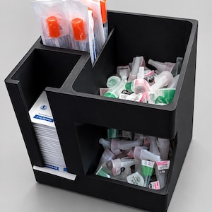 May include: A black multi-compartment organizer containing medical supplies. One section holds a stack of white paper packets labeled 2 Ply Medium, another holds orange-capped items in clear plastic, and two open bins contain various small medical tips.