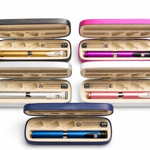 May include: A collection of colorful insulin pens in open cases. The pens are gold, silver, purple, pink, and blue. Each pen is accompanied by a clear vial. The cases are black, silver, pink, white, and blue.