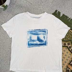 Pode incluir: Uma t-shirt branca de manga curta com uma impressão gráfica retangular centrada em tons de azul. O design apresenta uma representação artística e estilizada de uma zona do peito com texturas suaves e em espiral.
