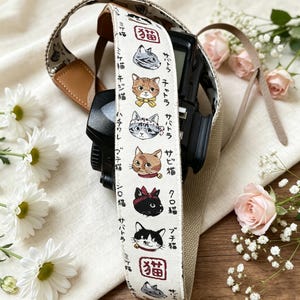 以下が含まれることがあります： クリーム色の布製バンドに様々な猫の顔の刺繍と日本語のテキストが施されたカメラストラップ。ストラップには茶色のレザーのアクセントがあり、黒いカメラ本体に取り付けられています。