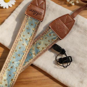 Peut inclure: Une courroie d'appareil photo en tissu bleu clair avec un motif d'étoiles dorées, des embouts en cuir marron et une sangle tissée marron. Un embout en cuir porte le nom Alice, l'autre un logo circulaire. Des clips en plastique noir sont fixés.