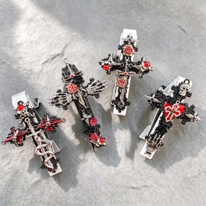 May include: Four silver-toned hair clips featuring cross-shaped designs. Each clip is adorned with intricate details including red and black roses, small red gemstones, skeletal hands, a coiled snake, a sword, and a small skull motif.