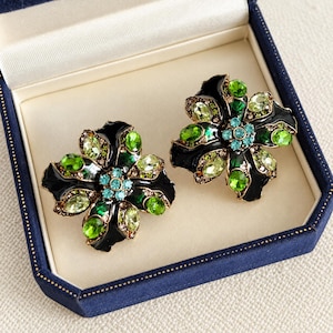 May include: A pair of flower-shaped earrings featuring black enamel petals accented with light green, dark green, and bright blue crystals. The center of each earring is adorned with a cluster of small blue stones set in a gold-toned metal frame.