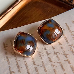 May include: A pair of square stud earrings featuring a polished, marbled resin surface with swirling patterns of deep amber and soft blue, encased in a delicate, beaded gold-tone metal frame with a reflective, glossy finish.