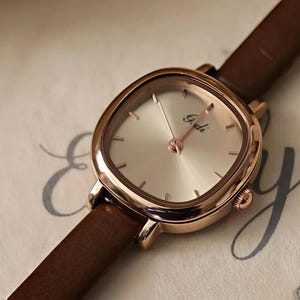 May include: A wristwatch with a rose gold-toned square case featuring rounded corners and a champagne-colored dial. The dial displays the brand name Gedi in black script and simple hour markers. It has a dark brown leather strap and a side crown.