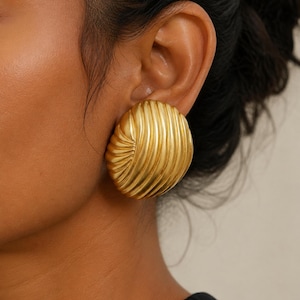 May include: A large, gold-toned earring with a rounded, shell-like shape featuring prominent, curved, vertical ridges. The surface has a matte, brushed finish, and the design is characterized by a swirling pattern that converges toward one side.