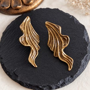 May include: A pair of gold-toned earrings featuring an elongated, abstract, and fluid wave-like design with deep, textured ridges. The metallic surface has a burnished finish, highlighting the organic, flowing curves of the metal shapes.