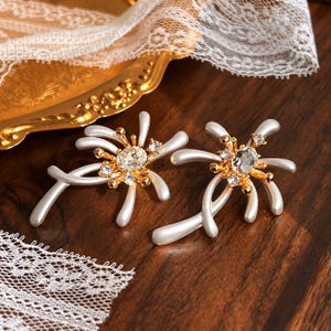 May include: A pair of gold-tone earrings featuring a central oval clear crystal surrounded by smaller round clear crystals and radiating white, smooth, curved, branch-like extensions that resemble a stylized floral or starburst shape.