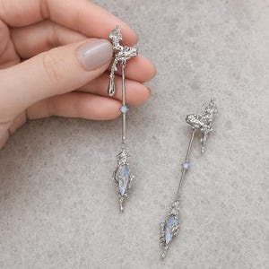 May include: A pair of silver-toned drop earrings featuring a textured, organic-shaped top piece, a long thin metal bar, and a central light blue oval stone encased in a jagged, metallic frame, with a small round blue accent bead on the bar.